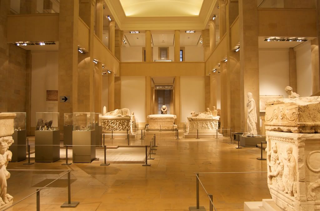 A photo of the National Museum of Beirut interior, displaying exhibits from different eras of Lebanese history, including ancient sculptures, pottery, and intricate gold jewelry from the Phoenician civilization.