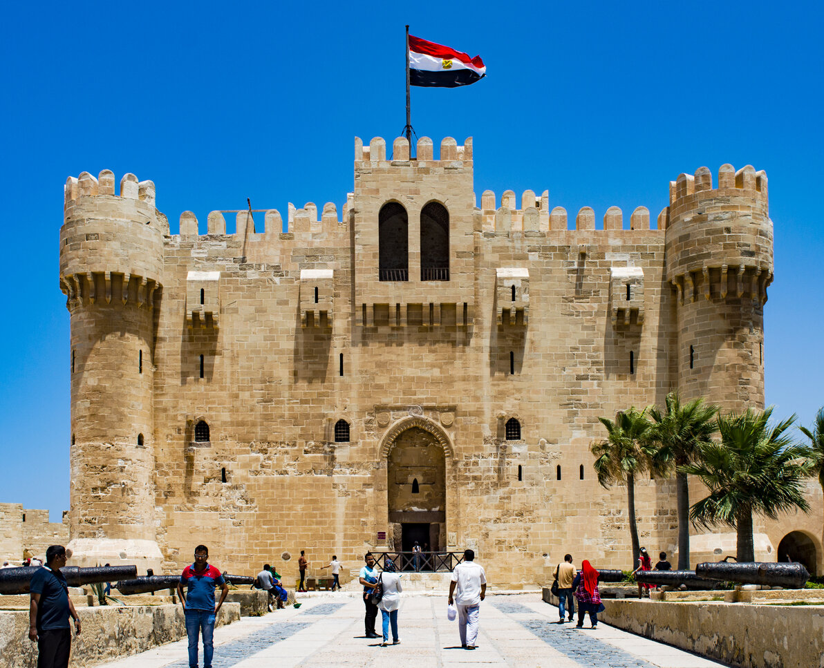 A towering stone fortress stands against the blue sky and overlooks the sparkling waters of the Mediterranean Sea. This is the Citadel of Qaitbay, a historic landmark in Alexandria, Egypt. The fortress has high walls, towers, and cannons that were used to defend the city against invaders. Visitors can explore the castle's various rooms, walk along the walls, and take in the stunning views of the sea. The Citadel of Qaitbay is a popular destination for tourists.