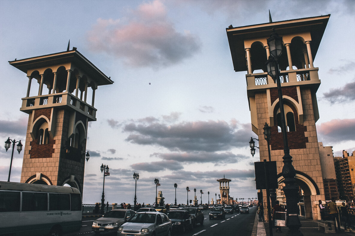 A beautiful panoramic view of Stanley Bridge, a white suspension bridge with four towering pillars and cars crossing over the sparkling blue water of the Mediterranean Sea. This is a popular landmark for tourists exploring things to do in Alexandria, Egypt.